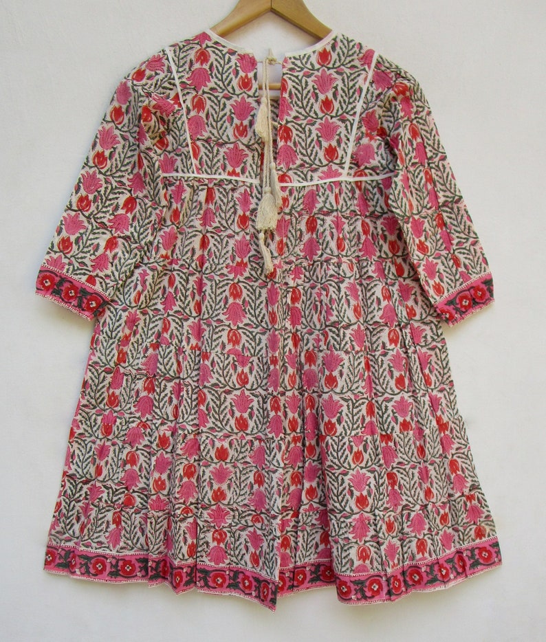 Puede incluir: Un vestido de algod&oacute;n blanco con estampado floral rosa y verde. El vestido tiene un escote redondo, mangas largas y una falda fruncida. El vestido se ata en la espalda con una borla blanca.