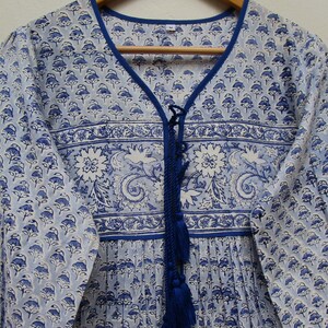 May include: A light blue and white floral print dress with a V-neckline and long sleeves. The dress features a blue trim, a decorative blue tassel, and a floral pattern. The dress is hanging on a wooden hanger.