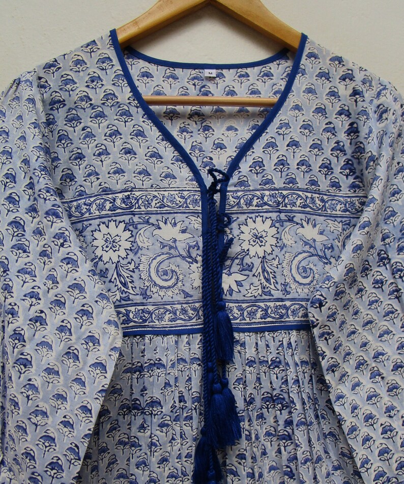 May include: A light blue and white floral print dress with a V-neckline and long sleeves. The dress features a blue trim, a decorative blue tassel, and a floral pattern. The dress is hanging on a wooden hanger.
