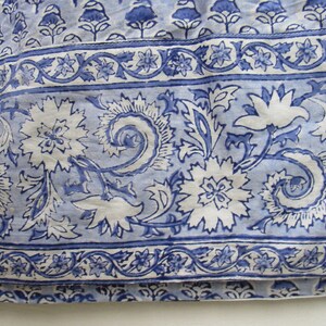 May include: Close-up of a blue and white floral patterned fabric. The design features large white flowers with intricate details, surrounded by blue leaves and swirls. A border with smaller floral and vine patterns is visible.