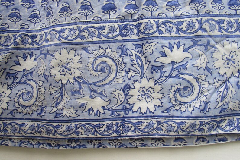 May include: Close-up of a blue and white floral patterned fabric. The design features large white flowers with intricate details, surrounded by blue leaves and swirls. A border with smaller floral and vine patterns is visible.