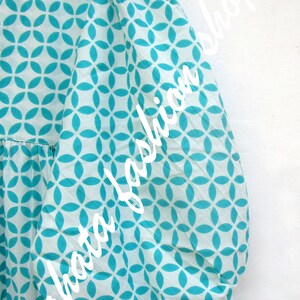 May include: A white dress with a turquoise geometric pattern. The dress has a ruffled collar and long sleeves with elastic cuffs.