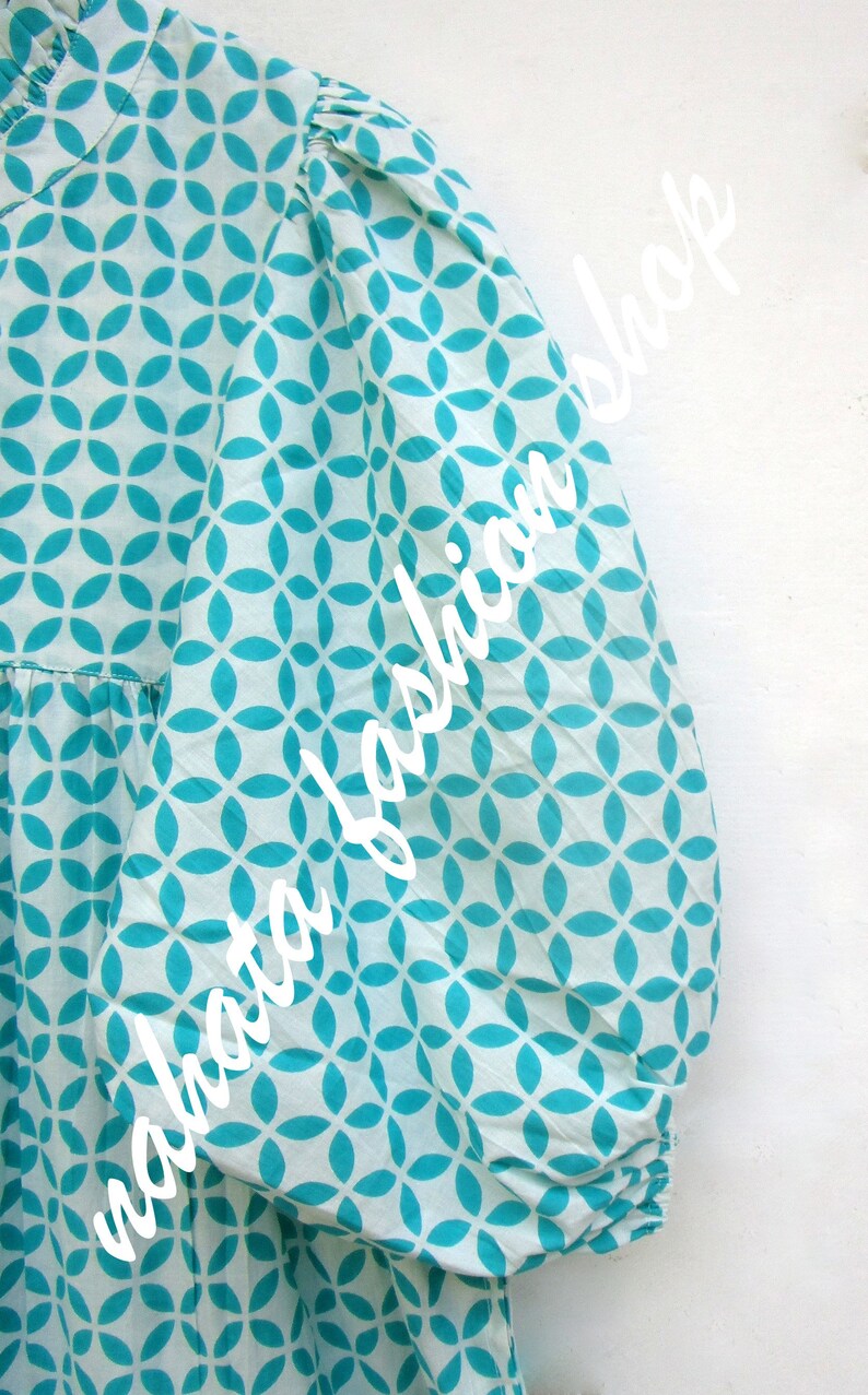 May include: A white dress with a turquoise geometric pattern. The dress has a ruffled collar and long sleeves with elastic cuffs.