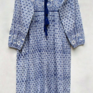 May include: A long, blue and white patterned dress with long sleeves and a V-neck. The dress features a floral and paisley design, with a blue tassel tie at the neckline. The dress is hanging on a wooden hanger.