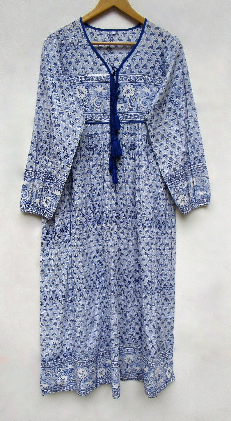 May include: A long, blue and white patterned dress with long sleeves and a V-neck. The dress features a floral and paisley design, with a blue tassel tie at the neckline. The dress is hanging on a wooden hanger.