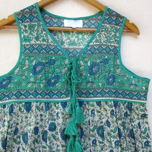 K&ouml;nnte beinhalten: Ein &auml;rmelloses Kleid mit gr&uuml;nem und wei&szlig;em Blumenmuster, einem Knoten am Ausschnitt und Quasten. Das Kleid hat einen wei&szlig;en Hintergrund mit einem gr&uuml;nen und blauen Blumenmuster.
