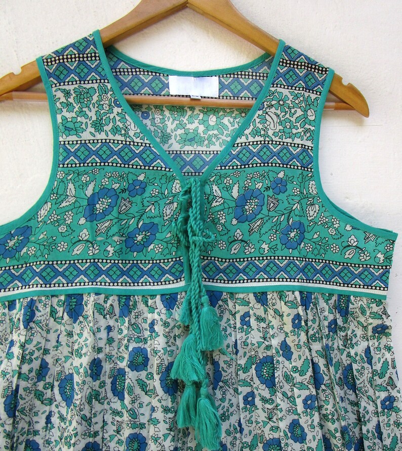 K&ouml;nnte beinhalten: Ein &auml;rmelloses Kleid mit gr&uuml;nem und wei&szlig;em Blumenmuster, einem Knoten am Ausschnitt und Quasten. Das Kleid hat einen wei&szlig;en Hintergrund mit einem gr&uuml;nen und blauen Blumenmuster.