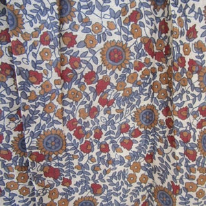 May include: Close-up of a fabric with a floral pattern. The design features sunflowers, small flowers, and leaves in shades of blue, red, and orange against a cream background. The fabric has a slightly textured appearance.