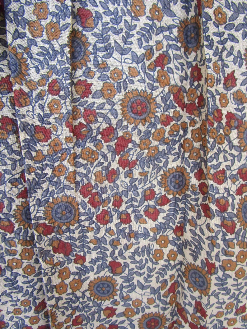 May include: Close-up of a fabric with a floral pattern. The design features sunflowers, small flowers, and leaves in shades of blue, red, and orange against a cream background. The fabric has a slightly textured appearance.