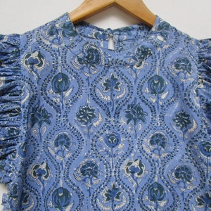 May include: A blue cotton top with a floral print. The top has a round neckline and short sleeves with ruffles.