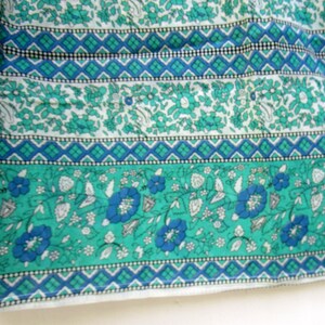 K&ouml;nnte beinhalten: Ein wei&szlig;er Stoff mit einem floralen Muster in Teal und Blau. Der Stoff hat ein sich wiederholendes Muster aus kleinen Blumen und Bl&auml;ttern. Der Stoff ist mit einem geometrischen Muster in Teal und Blau eingefasst.