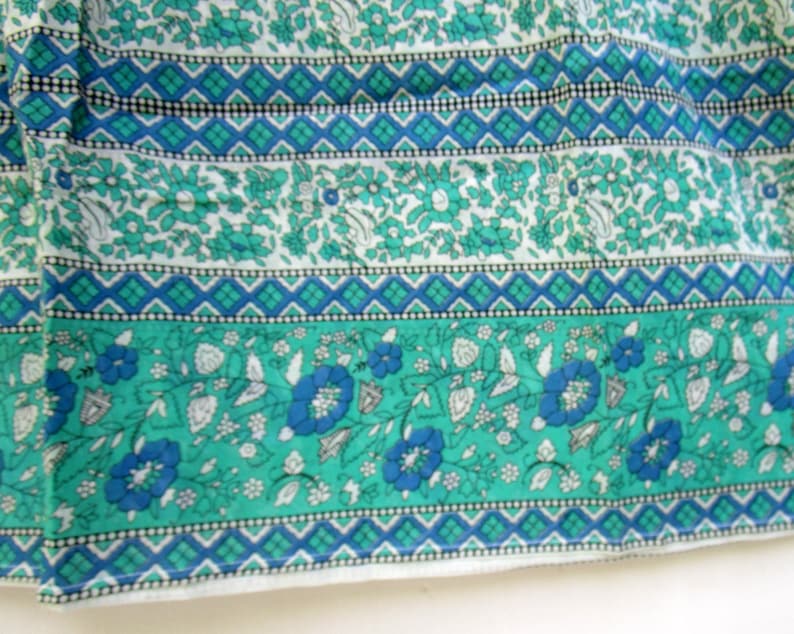 K&ouml;nnte beinhalten: Ein wei&szlig;er Stoff mit einem floralen Muster in Teal und Blau. Der Stoff hat ein sich wiederholendes Muster aus kleinen Blumen und Bl&auml;ttern. Der Stoff ist mit einem geometrischen Muster in Teal und Blau eingefasst.