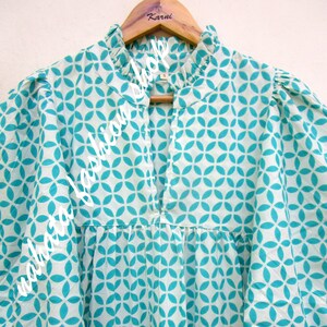 May include: A white cotton blouse with a teal geometric pattern. The blouse has a ruffled collar and a keyhole neckline.