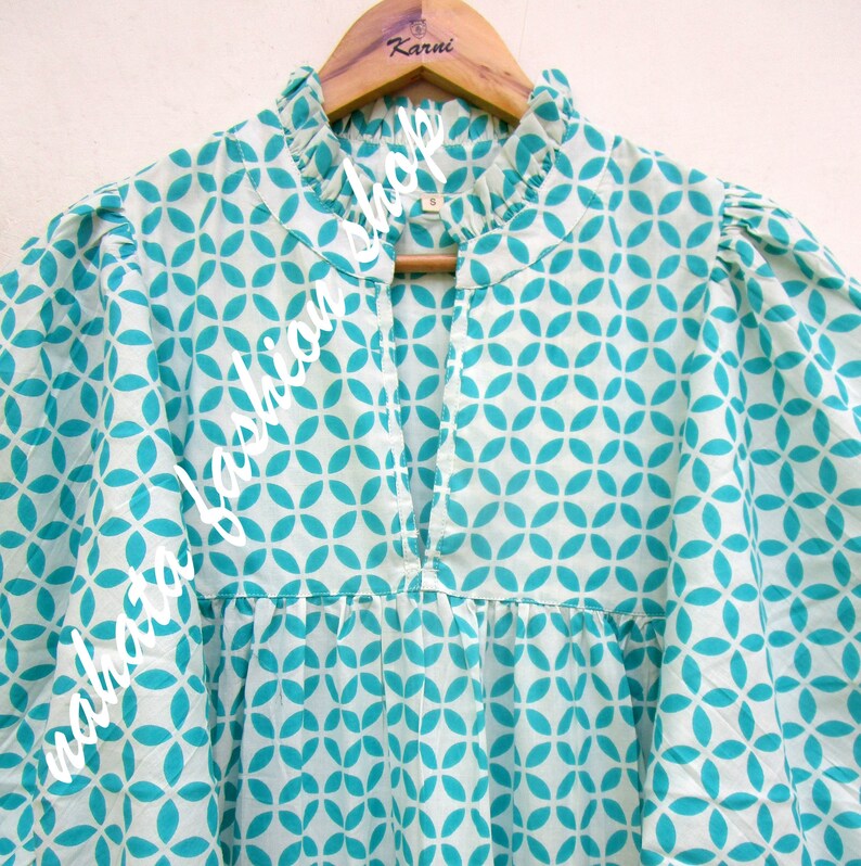 May include: A white cotton blouse with a teal geometric pattern. The blouse has a ruffled collar and a keyhole neckline.