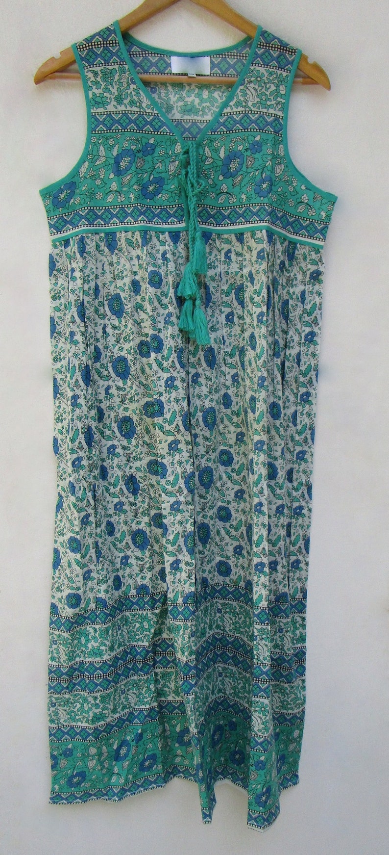 K&ouml;nnte beinhalten: Ein &auml;rmelloses Kleid mit einem Blumendruck in Blau- und Gr&uuml;nt&ouml;nen. Das Kleid hat einen Knoten am Ausschnitt und ein Quasten-Detail.