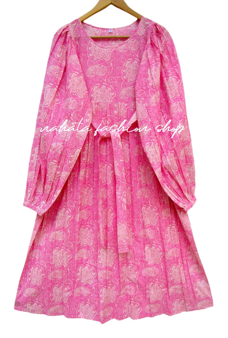 May include: A pink dress with a paisley pattern and long sleeves. The dress has a tie at the waist and the text "nahata fashion shop" is visible. The dress is hanging on a wooden hanger.