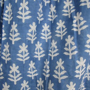 May include: Close-up of a blue fabric with a white floral pattern. The pattern features stylized white flowers and leaves arranged in a repeating design. The fabric has a slightly textured appearance.