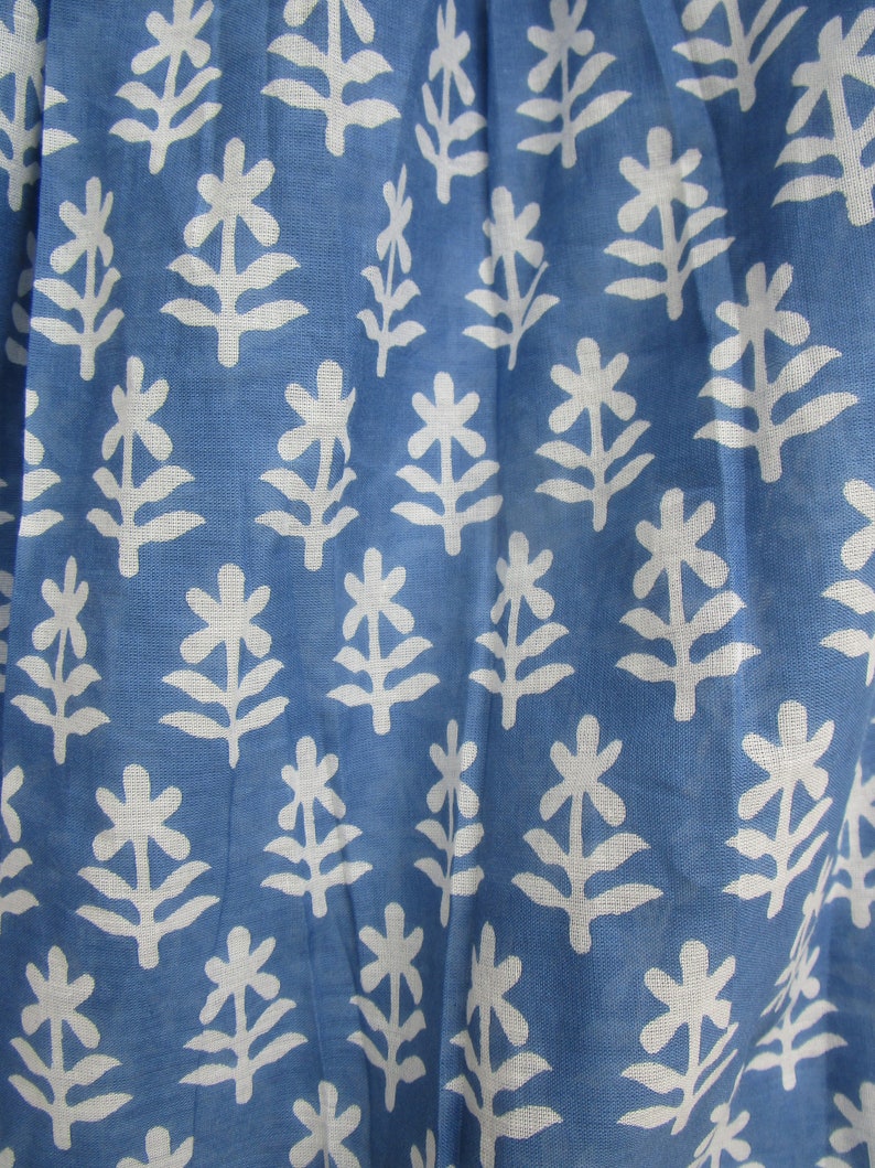 May include: Close-up of a blue fabric with a white floral pattern. The pattern features stylized white flowers and leaves arranged in a repeating design. The fabric has a slightly textured appearance.