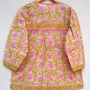 May include: A yellow and pink floral print cotton tunic top with long sleeves and a round neckline. The top has a pink trim around the neckline and a yellow trim around the sleeves and hem.