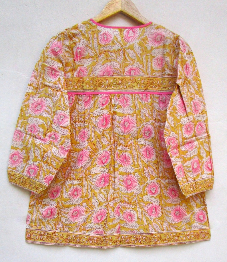 May include: A yellow and pink floral print cotton tunic top with long sleeves and a round neckline. The top has a pink trim around the neckline and a yellow trim around the sleeves and hem.