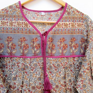 May include: A floral print tunic top with a V-neckline and a tassel tie. The top features a gray band with orange and red floral designs, accented with a bright pink trim. The fabric has a beige background with a repeating floral pattern.