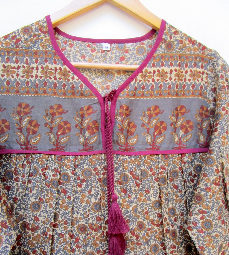 May include: A floral print tunic top with a V-neckline and a tassel tie. The top features a gray band with orange and red floral designs, accented with a bright pink trim. The fabric has a beige background with a repeating floral pattern.