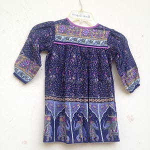 May include: A navy blue dress with a floral pattern and elephant motifs. The dress features long sleeves, a round neckline, and a decorative border. The dress is hanging on a white hanger.