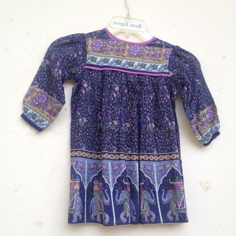 May include: A navy blue dress with a floral pattern and elephant motifs. The dress features long sleeves, a round neckline, and a decorative border. The dress is hanging on a white hanger.