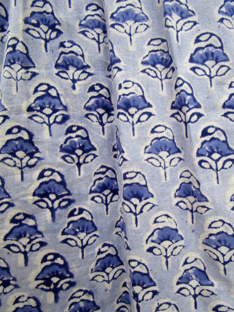May include: Close-up of a light blue fabric with a repeating pattern of dark blue floral motifs. The flowers have a stylized design with rounded petals and stems. The fabric is slightly wrinkled, suggesting it is a soft material.