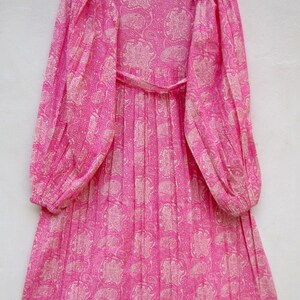 May include: A long-sleeved, pink dress with a paisley pattern. The dress features a V-neck, a cinched waist with a matching belt, and pleated skirt. The sleeves are long and gathered at the cuffs. The dress is hanging on a wooden hanger.