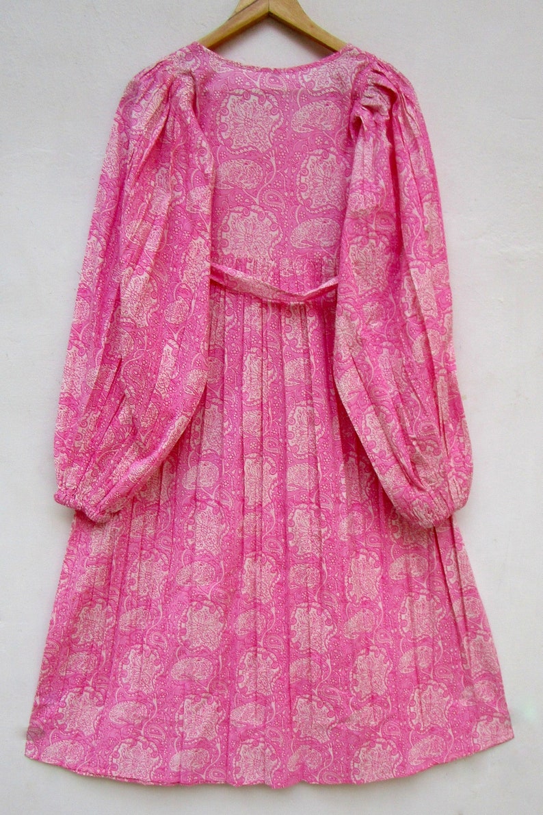 May include: A long-sleeved, pink dress with a paisley pattern. The dress features a V-neck, a cinched waist with a matching belt, and pleated skirt. The sleeves are long and gathered at the cuffs. The dress is hanging on a wooden hanger.
