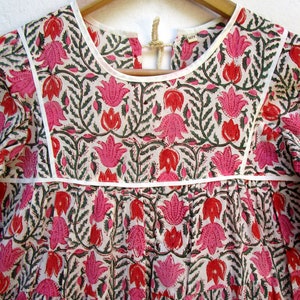 Puede incluir: Una blusa de algod&oacute;n blanca con estampado floral rosa y verde. La blusa tiene un escote redondo y mangas cortas. La blusa est&aacute; en una percha.
