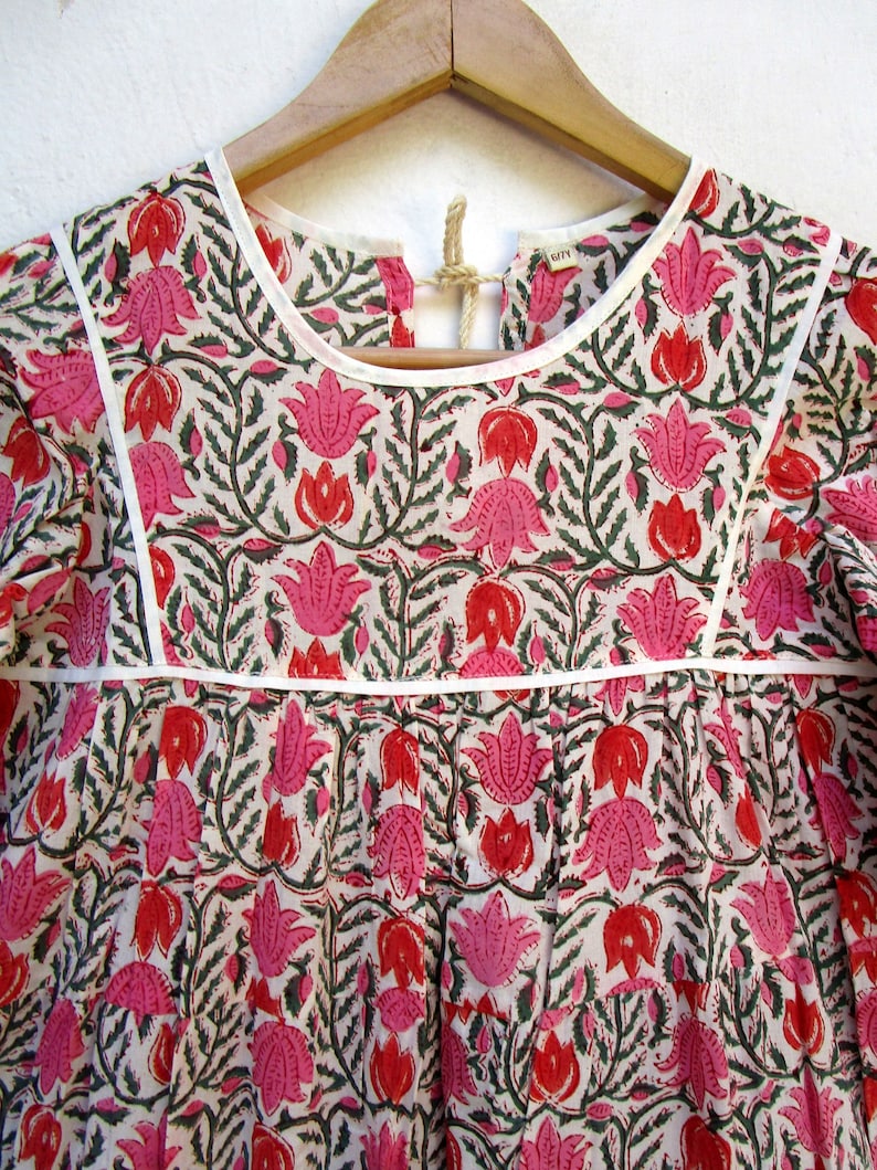 Puede incluir: Una blusa de algod&oacute;n blanca con estampado floral rosa y verde. La blusa tiene un escote redondo y mangas cortas. La blusa est&aacute; en una percha.