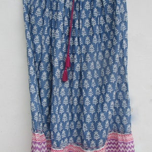 May include: A blue skirt with a white floral pattern and a pink and purple chevron border. The skirt has a drawstring waist and a pink tassel. The skirt is hanging on a wooden hanger.