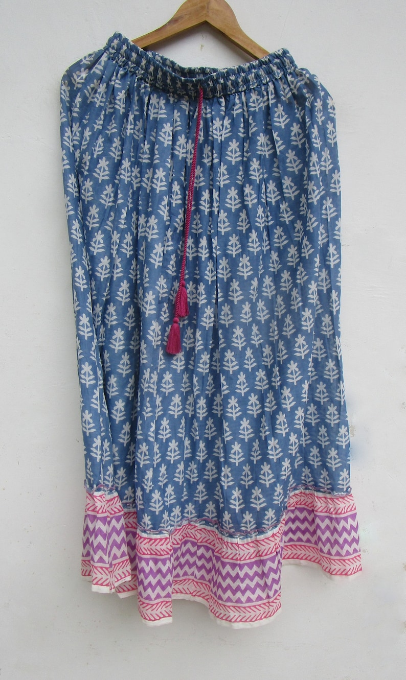 May include: A blue skirt with a white floral pattern and a pink and purple chevron border. The skirt has a drawstring waist and a pink tassel. The skirt is hanging on a wooden hanger.