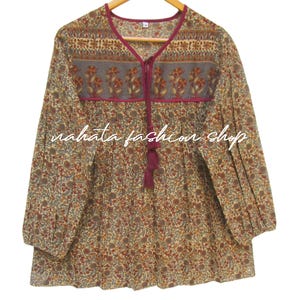 May include: A long-sleeved blouse with a floral pattern in shades of brown, red, and beige. The neckline and trim are a deep burgundy, with a matching tassel. The text "nahata fashion shop" is printed on the front.