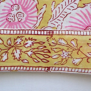 May include: Close-up of a yellow fabric with a pink floral pattern and a red and white border. The fabric has a repeating pattern of flowers and leaves.