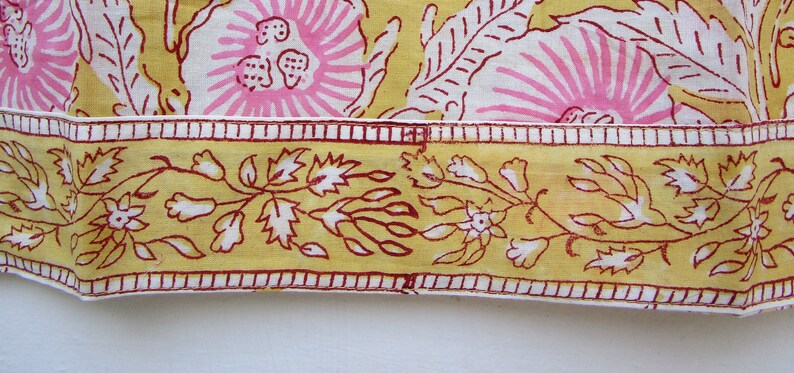 May include: Close-up of a yellow fabric with a pink floral pattern and a red and white border. The fabric has a repeating pattern of flowers and leaves.