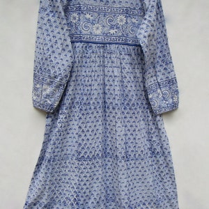 May include: A long, light blue dress with a floral pattern and long sleeves. The dress features a round neckline and a gathered waist. The fabric is adorned with a repeating blue and white floral design, with a decorative border at the hem and sleeves.