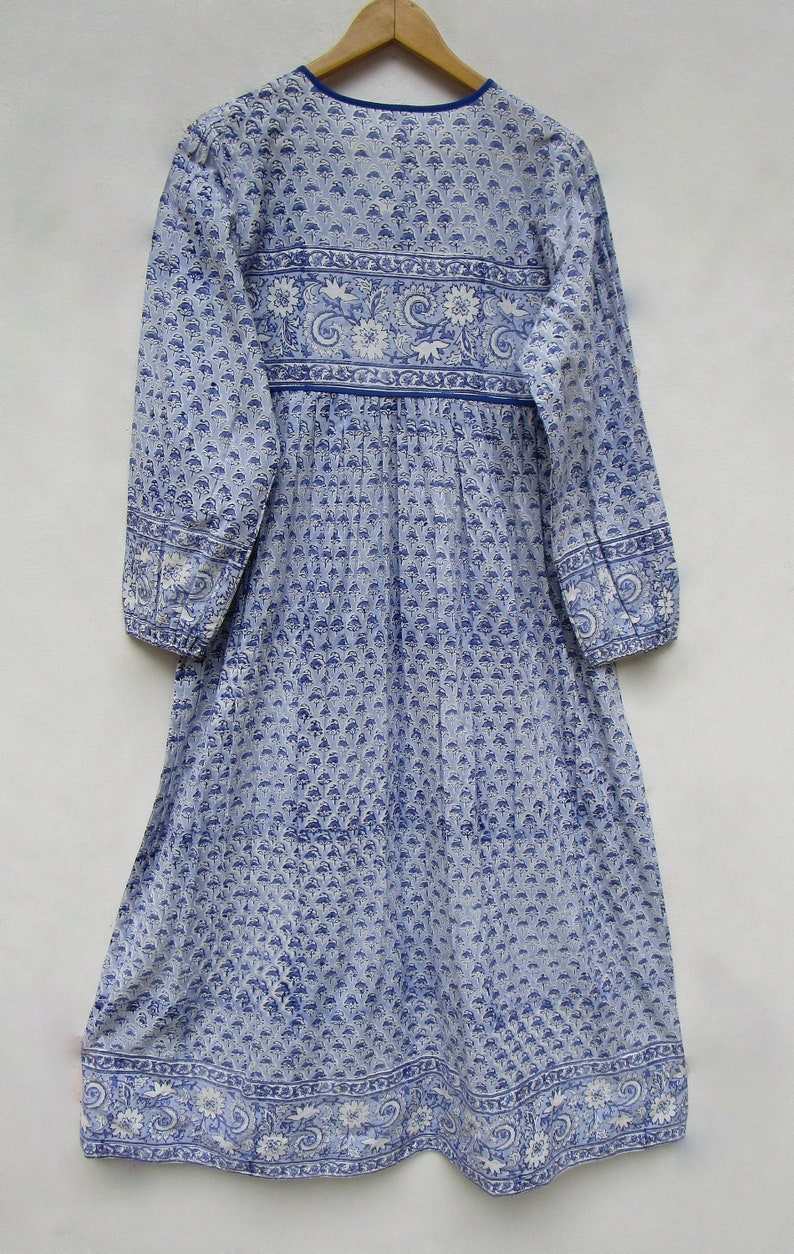May include: A long, light blue dress with a floral pattern and long sleeves. The dress features a round neckline and a gathered waist. The fabric is adorned with a repeating blue and white floral design, with a decorative border at the hem and sleeves.