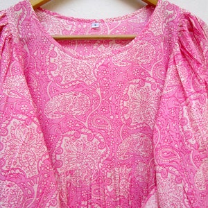 May include: A pink dress with a white paisley pattern, hanging on a wooden hanger. The dress has a round neckline, puffed sleeves, and a tie belt. The fabric is lightweight and appears to be cotton or a similar material.