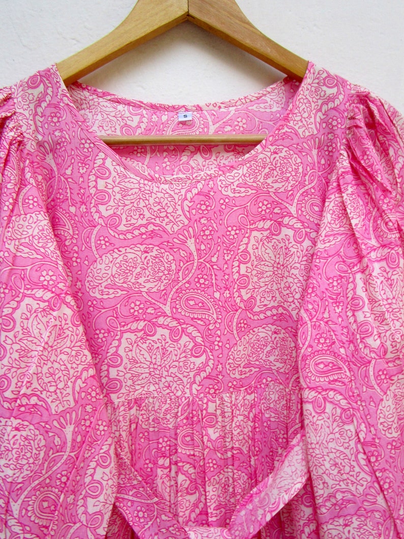 May include: A pink dress with a white paisley pattern, hanging on a wooden hanger. The dress has a round neckline, puffed sleeves, and a tie belt. The fabric is lightweight and appears to be cotton or a similar material.