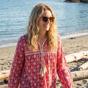 Puede incluir: Una blusa roja de manga larga con estampado floral, con cuello en V y lazo con borlas. La blusa presenta un borde color crema y un patrón floral repetido. La persona de la imagen lleva gafas de sol y está en la playa.