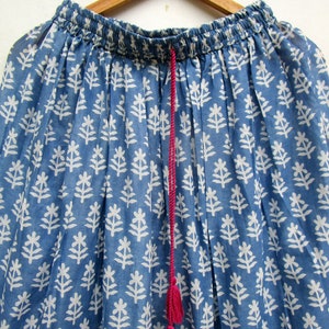 May include: A blue skirt with a white floral pattern, hanging on a wooden hanger. The skirt has an elasticated waistband and a pink drawstring with tassels. The fabric is lightweight and appears to be cotton.