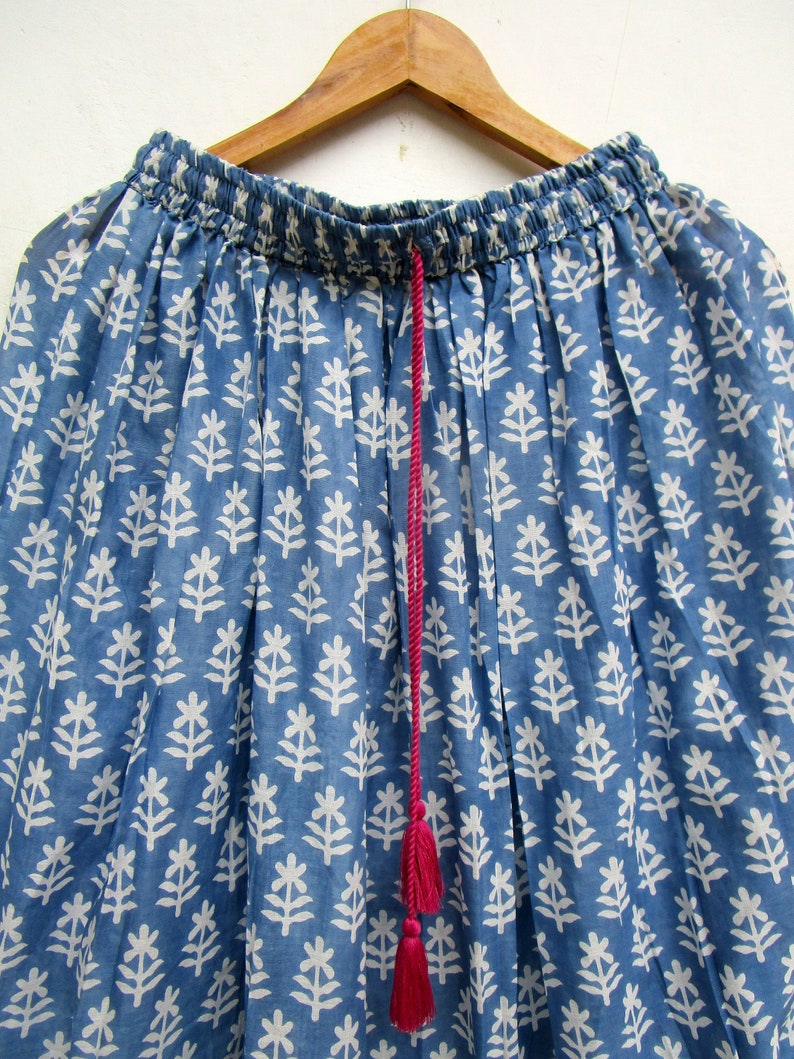 May include: A blue skirt with a white floral pattern, hanging on a wooden hanger. The skirt has an elasticated waistband and a pink drawstring with tassels. The fabric is lightweight and appears to be cotton.