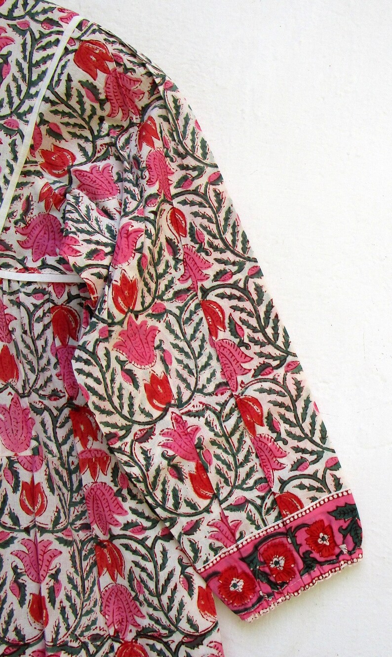 Puede incluir: Una blusa de algod&oacute;n blanco con estampado floral rosa y verde. La blusa tiene manga larga y un pu&ntilde;o fruncido. La tela es ligera y tiene una textura suave.