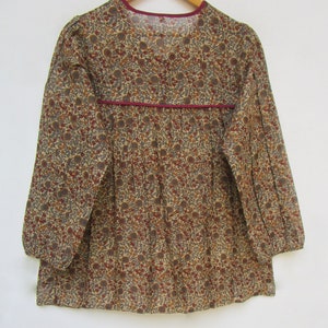 May include: A long-sleeved blouse with a floral pattern in shades of brown, red, and gray on a beige background. The blouse has a V-neckline and a burgundy trim along the neckline and waist. The sleeves are gathered at the cuffs.
