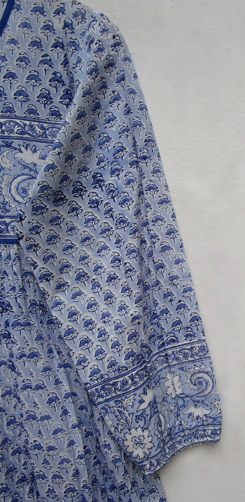 May include: A light blue and white patterned garment with a floral design. The fabric features a repeating pattern of blue flowers and a decorative border along the sleeve and neckline. The garment appears to be a long-sleeved top or dress.
