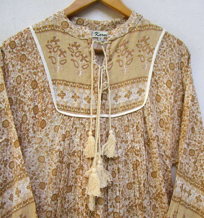 May include: A white and brown floral print cotton tunic top with a tie neckline and tassel details. The top has a white trim around the neckline and a small label that says "Karuni" on the front.