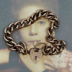 May include: A gold heart-shaped padlock charm bracelet with a curb chain. The bracelet is on a vintage photograph of a young girl.